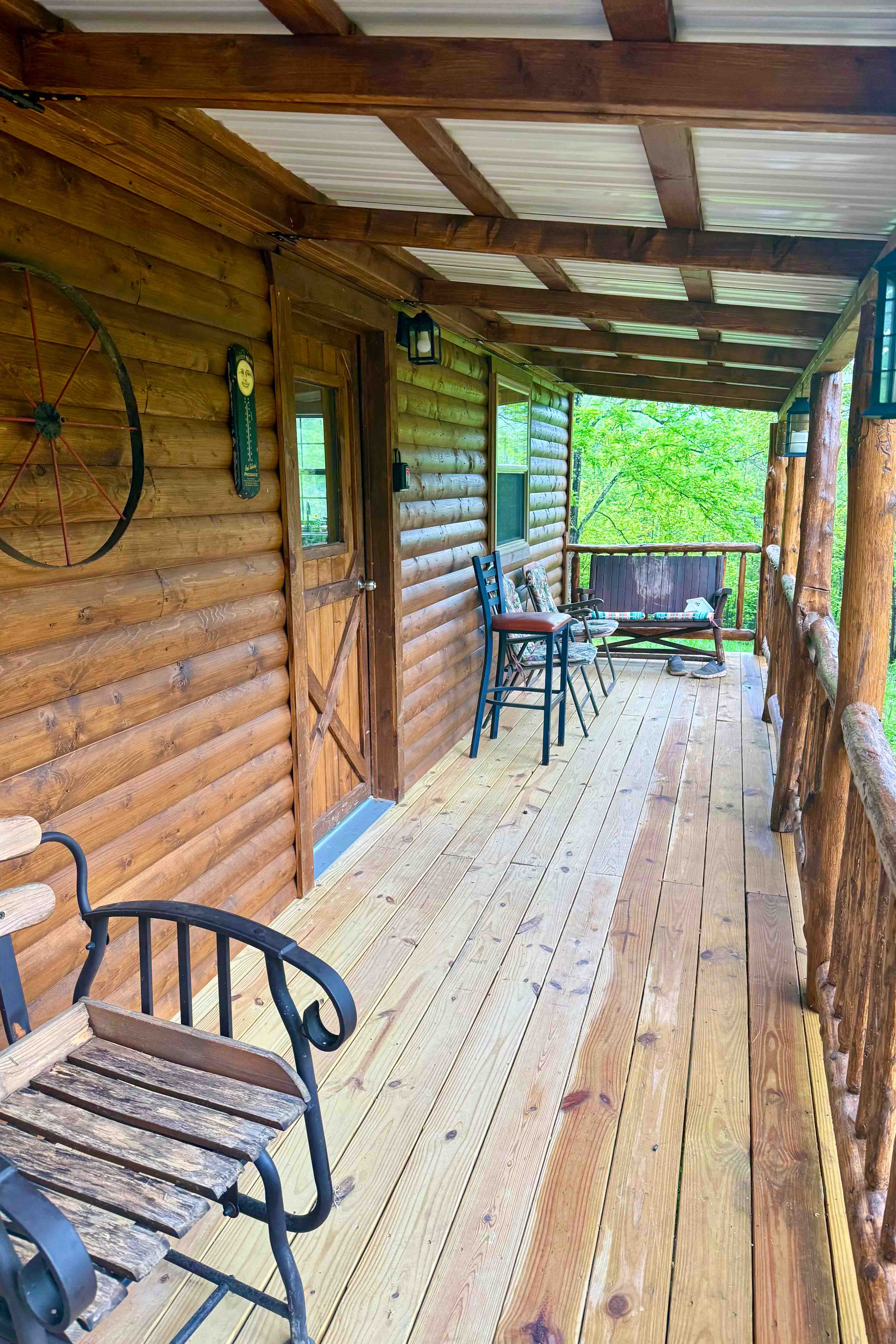 A view from the front porch of our newest mountain cabin. The porch is rustic with wooden chairs and a wagon wheel on mounted on the wall. Beautiful green trees can be seen in the background.